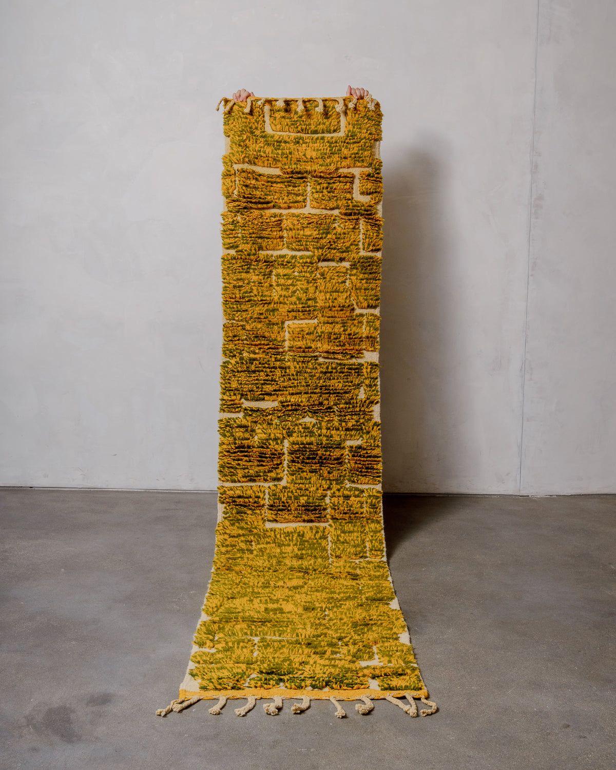 Berberlin Berber runner rug in mixed yellow and olive green wool, being held up in front of a white wall.